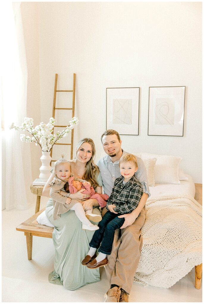 family of 5 sitting on a white bed posing for newborn photos in a photography studio