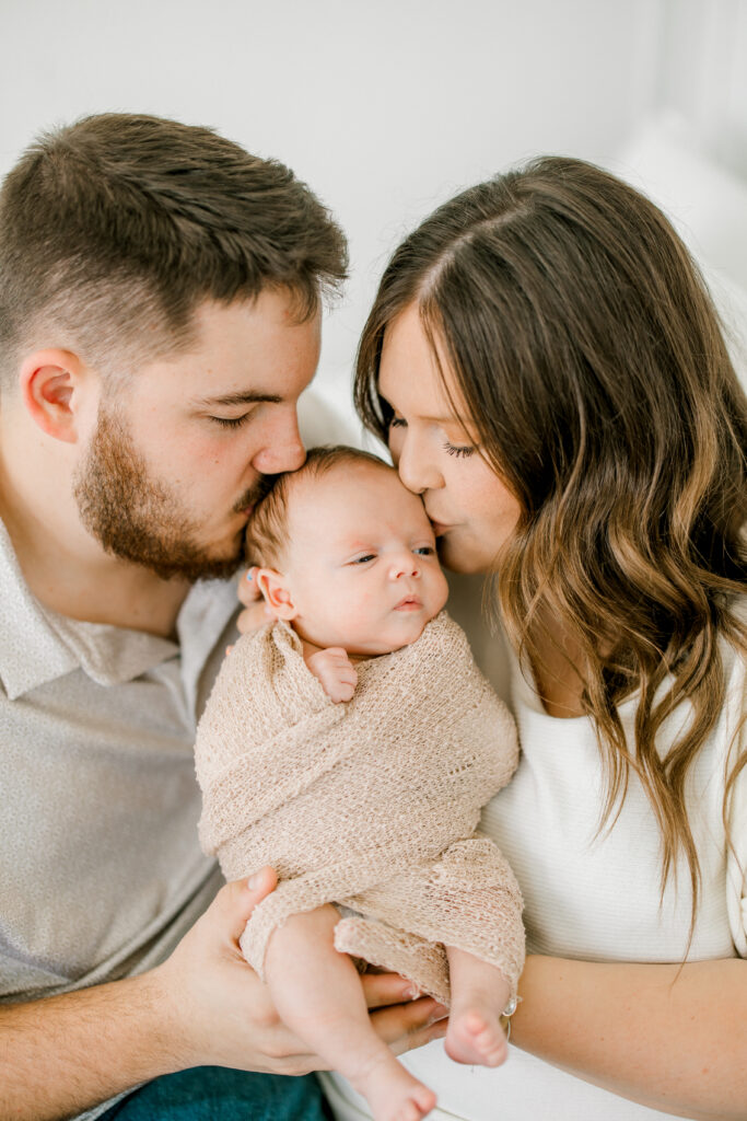 Parents kissing newborn Baby Beau during indoor studio session